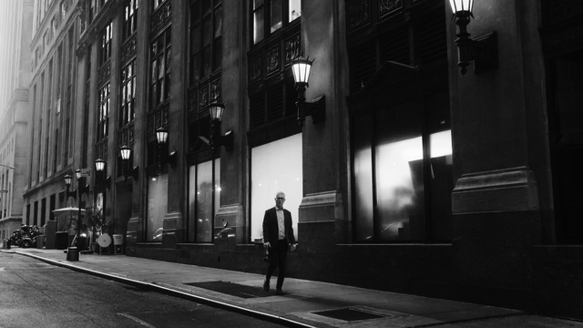 A person in a suit walks in front of a bright window of a large gothic building. On the building are light fixtures.