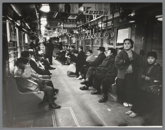 The image depicts a scene inside what appears to be a subway or metro train. A group of people are seated and standing in the aisle, facing opposite directions as if they were going their separate ways after work hours.

On one side, there is a row of passengers sitting close together; some appear tired or weary, while others engage with each other through conversation. The individuals vary from men to women, all dressed in what seems like casual attire typical for the time period suggested by the image's black and white quality—mid-20th century clothing styles.

On the opposite side of the aisle, a few more people are standing or sitting on benches that line this part of the train. One woman is notably bent over, holding onto something in her hands, perhaps waiting for someone to join her or checking what she holds closely.

The interior has circular metal rings hanging from above and various signs with Japanese characters indicating it's likely a photograph taken in Japan during 1960s as mentioned by the metadata. The lighting is subdued, typical of public transportation interiors at night time. There are no prominent features like advertisements or modern amenities that might suggest this was recently captured.

The overall atmosphere conveyed through body language and arrangement suggests daily routine and perhaps fatigue after a day's work for most passengers.