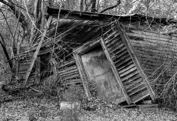 A black and white photo of an abandoned wooden structure which has partially collapsed on the left causing a severe tilt in that direction.