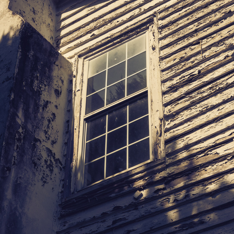 A close-up view of a deteriorating double-hung window on an abandoned building, shot from below at an angle. The window has white painted wooden frames with multiple panes in a grid pattern. The paint on both the window frame and the horizontal wooden siding is severely peeling and flaking, exposing bare, weathered wood underneath. The siding runs diagonally across the frame due to the camera angle. On the left side, there's a corner or column with heavily chipped and peeling paint showing layers of white and darker colors beneath. The window glass appears dirty and reflects some shadows or interior darkness. The overall image has a sepia or vintage tone, emphasizing the aged and neglected condition of the structure.