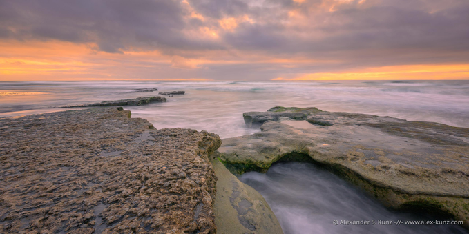 A wide format color seascape photo showing a mostly overcast sky above rock formations that are surrounded by rushing waves. The scene is tinted in the warm colors of sunset and the motion of the water is blurred by a slow shutter speed.