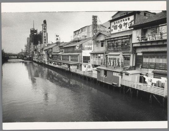 The image is a black and white photograph depicting an urban riverside scene, likely from the mid-20th century. A calm river runs parallel to numerous buildings with distinct East Asian architectural features such as tiled roofs, wooden balconies, and multi-storied structures close together along its edge. The area appears densely populated or developed for commercial use given the various signs in a non-Latin script affixed above some of the windows.

The water reflects the outlines of the surrounding architecture and adds to the tranquil atmosphere of the scene despite the bustling urban setting suggested by the buildings' arrangement and density. On closer inspection, clothes appear hung on lines outside one structure's balcony, indicating domestic life amid commercial activity. There are no visible people or moving vehicles that can be discerned from this perspective.

The photograph is a historical record capturing the essence of Osaka’s riverside architecture in 1960 as documented by Ed van der Elsken, noted for his work on street photography and cityscapes during post-war Japan's economic boom. The image serves not only to provide a visual experience but also offers insights into urban living conditions, commercialism along waterfronts, and the architectural styles of that era in Osaka.