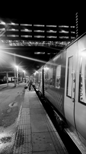 Looking along the length of a train and platform at night, above and in the distance a large building on the road above the station