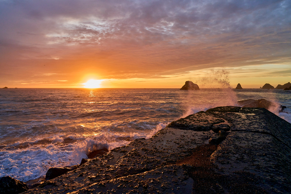 A concrete jetty extends toward a few off shore sea stacks. The sky is ablaze in oranges and pinks, and the light is reflected on the water covering the jetty. There are waves breaking against the jetty. In the distance the sun is about to drop below the horizon.