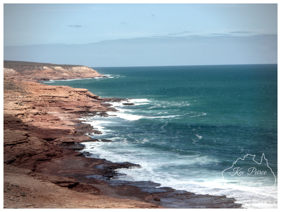 A sweeping panoramic landscape of the rugged Kalbarri coastline in Western Australia. Red and brown layered sandstone cliffs drop sharply to the deep turquoise ocean, where white waves crash onto the shore.

The horizon is flat under a pale blue sky. The signature 'Kev Peirce' is visible in the bottom left corner.