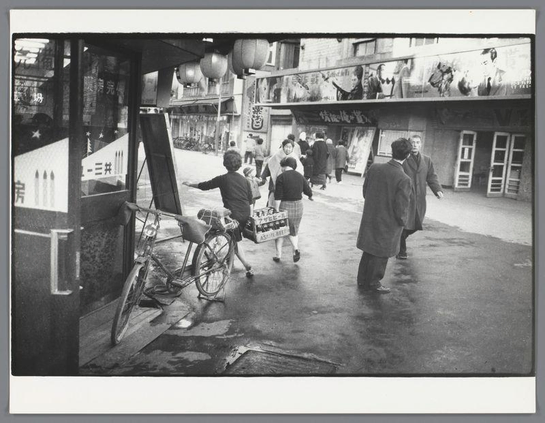 The image depicts a bustling street scene captured in black and white. Several individuals are navigating the sidewalk, including two women carrying boxes of beer as they walk away from the camera's viewpoint. One woman is extending her arm out to balance one end while the other follows closely behind with their box resting on top of what appears to be another bicycle or wheeled vehicle that has tipped over.

The street environment includes various signs and advertisements in Japanese, indicating a commercial area likely situated in Japan during mid-20th century. The setting suggests an urban landscape with shops lining one side of the narrow alleyway. Overhead, hanging lanterns add to the ambiance of this busy thoroughfare.

One prominent figure is walking on the right-hand path away from the camera, wearing a coat and looking ahead as if engaged in conversation or moving towards their destination. Other pedestrians are also visible around them; some appear to be conversing with one another while others simply walk by without interaction.

The ground shows signs of wear and possible water damage, which could suggest either recent rain or poor drainage conditions typical for urban areas during the era captured in this photograph. The overall impression is that of a vibrant street life within an East Asian cityscape from several decades ago.