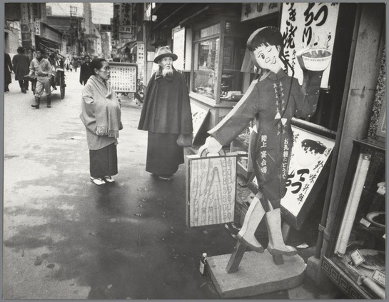 The image captures a lively street scene in what appears to be Japan, possibly Osaka. Two individuals are engaged with an advertisement board featuring the animated character from "Nausicaä of the Valley of the Wind." The larger figure is dressed as Captain Jean Dufort and holds out a price list or menu for passersby to view.

In the background, various people navigate through what seems like a commercial district. Some are walking past shops with storefronts displaying signs in Japanese characters. A person on the left side of the frame wears traditional Japanese attire, indicating cultural preservation amidst urban life. The image has an air of candid street photography, capturing everyday moments and local culture.

The photograph is presented in black and white, which suggests it might be from a past era or intended to evoke that vintage aesthetic. Overall, this scene illustrates contemporary daily life with touches of traditional Japanese elements alongside modern influences introduced by the animated character advertisement board.