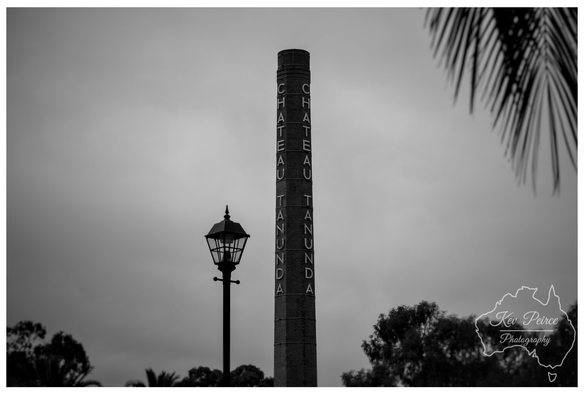 Full colour photograph of the main facade of Chateau Tanunda, featuring symmetrical red brick and stone work, arched windows, and dark sloping roofs.  A wide, bright green lawn and a central gravel pathway lead the eye toward the grand entrance under a bright blue sky with wispy white clouds.