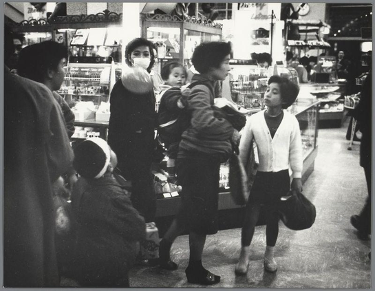 The image depicts a scene inside what appears to be an Asian supermarket. A group of people, possibly East Asian based on their features and clothing styles, are engaged in various activities typical of shopping: browsing items, conversing with each other or store staff, and attending to personal needs such as handling bags or adjusting accessories like scarves.

In the center-right of the image stands a woman wearing glasses, likely spectacles given her age. She seems attentive or perhaps waiting for something; she is dressed in what looks like business casual attire—a white cardigan over a darker top paired with trousers and shoes suitable for walking. Her posture suggests engagement but also readiness to move.

To her left, another older individual appears deeply engrossed in examining items on the shelves, possibly reading labels or considering products they might purchase. They are wearing glasses, suggesting vision impairment which is common among aging populations. The person's attire includes a dark-colored coat and hat, indicating that it could be cold weather outside.

On her right side stands another woman holding a child close to herself. She wears what seems like traditional Japanese clothing—a white top with an apron-like skirt—commonly worn by shopkeepers or housewives in Japan for practical reasons while shopping. The child appears content and relaxed, indicating they are comfortable within this setting w [...]