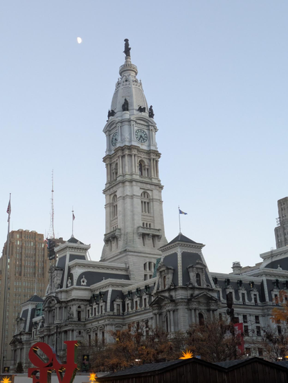 Photograph facing Philadelphia's city hall, which has a tall central tower, topped by a large statue of William Penn. The moon appears to the left of that statue. In the lower left of the scene, most of the block sculpture, "LOVE," by artist Robert Indiana, can be seen. The bottom of the letters "v" and "e" are not in the frame. All of the letters are backwards because the sculpture is seen from behind. It is in Love Park, across the street from City Hall.