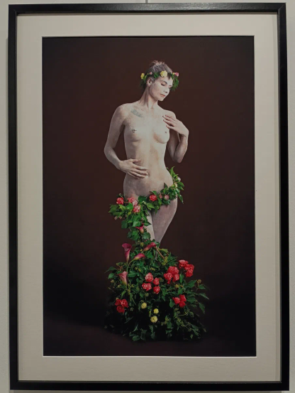 Photographie encadrée montrant une femme nue posant debout, le corps partiellement couvert de feuillage et de fleurs colorées, avec une couronne florale sur la tête, sur fond uni sombre.