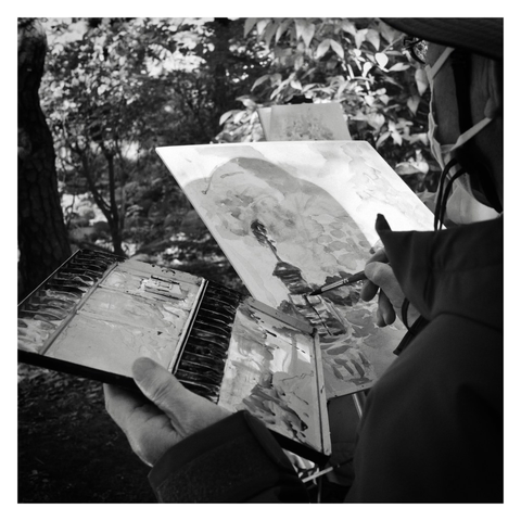 Black and white photo close-up of an artist painting an outdoor scene.
