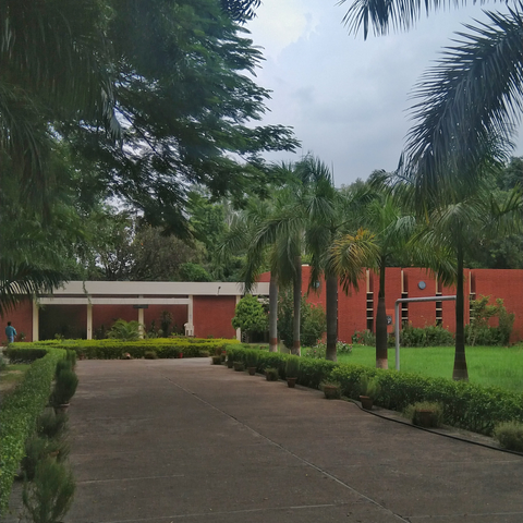 A red brick building is surrounded by lush greenery and tall palm trees under a cloudy sky. A wide walkway leads to the entrance, lined with trimmed bushes.