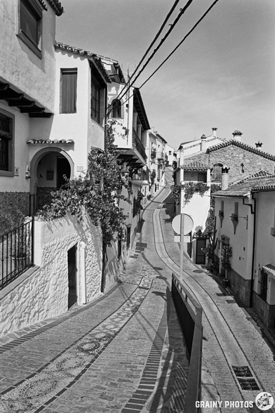 A narrow cobblestone street winds through quaint white houses with tiled roofs, framed by lush greenery. The scene, captured in black and white, evokes a charming atmosphere of a picturesque hillside Spanish village.