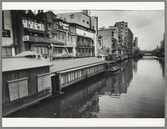 The image depicts a black and white street scene along a canal, likely taken in the early to mid-20th century. A series of multi-story buildings line one side of the waterway with signage in Japanese characters visible on several facades, indicating businesses or residences. The architecture suggests an urban setting, possibly from Japan given the script.

The foreground features wooden structures that could be warehouses or residential extensions overhanging the canal, adding to a sense of layered use of space along the waterfront. A small boat is moored at one end of these extensions on the waterway. Reflections and ripples in the calm surface hint at gentle movement within the water.

The overall atmosphere conveyed by the image suggests an everyday scene from life beside a river or canal, emphasizing the interaction between urban living spaces and natural elements like waterways.