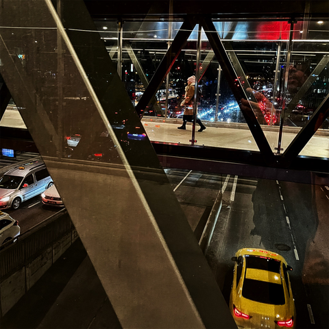 A pedestrian walks on a glass walkway above a busy street, with a yellow taxi and city lights visible below at night.