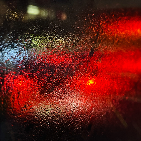 A close-up of a wet windshield showing blurred red and yellow lights, creating a colorful, abstract, and atmospheric effect.