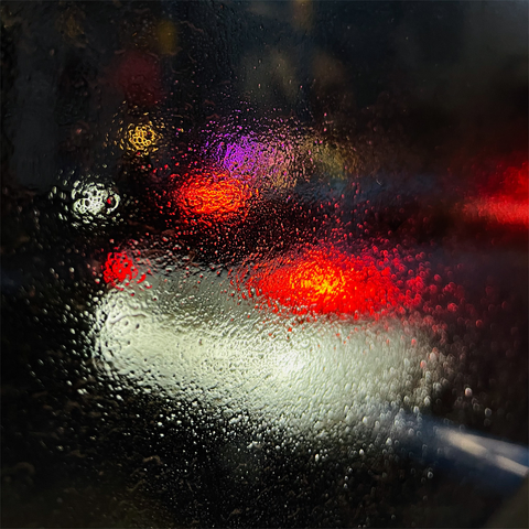 A blurred, raindrop-speckled window reveals bright red and white lights from vehicles passing by in a nighttime urban setting.