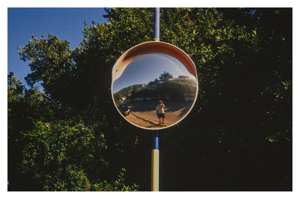 Color photograph of a round traffic mirror framed in yellow, standing in front of a few green trees. In the mirror, you can see the photographer holding the camera in one hand and a dog leash in the other. Unfortunately, the dog is not visible. Behind the photographer in the mirror image, you can see residential buildings and a parked car on the left.