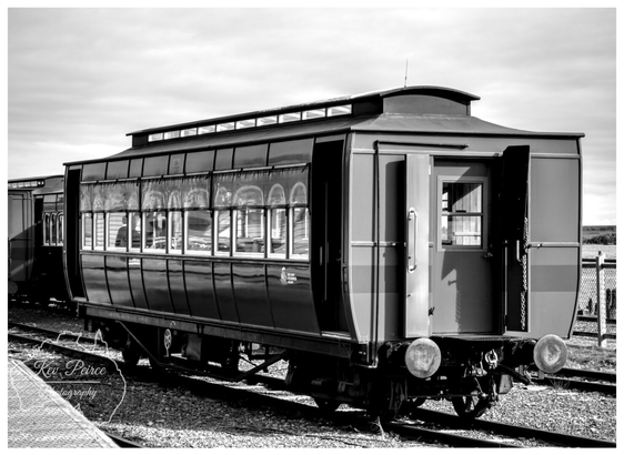 A striking black and white photograph of an old, preserved passenger train carriage, possibly from the Abt Railway, sitting on the tracks at Strahan, Tasmania.  The carriage features large windows along its side and open doors at the end. The image, signed Kev Peirce Photography, captures the vintage rail heritage under an overcast sky.