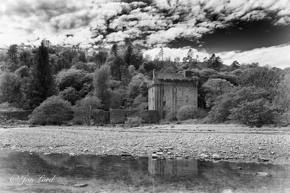This is a black and white photo in landscape format of a restored Scotish castle in a rural setting. Saddell, Kintyre, Scotland (2017).

In the foreground and stretching across the base of the image and rising upwards less that a quarter up the photo are the still and clear waters of a small stream that is flowing slowly in to the sea that is out of view behind the camera. Beyond is a stony and fairly flat beach that rises up to the one-third point in the image. There and slightly to the right of centre is a small castle, that is square, between ten and fifteen metres along each wall: infact almost a cube. The walls are light grey with some weathered dark streaks running down the walls. Set in these walls are some randomly placed and sized, white, rectangular windows, six on the wall facing the camera and four on the wall to the left. On the top of the sides of the castle is a castillated wall with defensive notches spaced every few metres along its length. Behind this wall is a pitched and tiled roof with a large chimney on the right end and a smaller chimney at the left end. To the immediate left of the castle is a derelict, single story and roofless stone building. In the background is the rising terrain of a forrested ridge that stretches across the image from left to right. Above is a partly cloudy sky with medium level clouds and clear patches to the right. 

This is Saddell Castle that was once one of the ancestrial homes of the MacDonald clan in Scotland.
