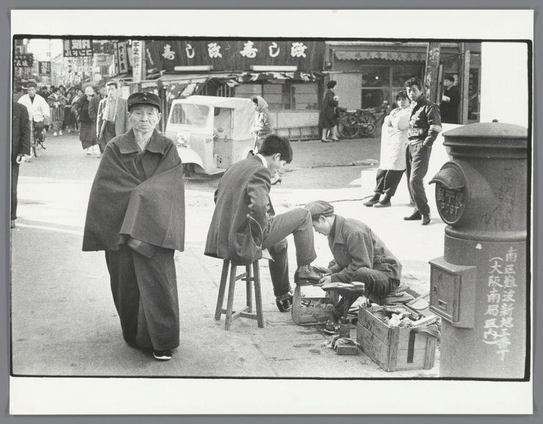 The image depicts a street scene with multiple individuals engaged in various activities. In the foreground, on the left side of the frame, there is an older man draped in what appears to be a shawl or cloak, looking off into the distance and seemingly observing his surroundings. He stands out due to his size compared to others around him.

Moving towards the center, two younger men are seated; one sits on a stool while he seems to be examining something with concentration, possibly paperwork or tools of some sort, and another is kneeling beside an open suitcase filled with various items, likely belonging to them. Their attire suggests they might be shoeshine workers.

On the right side of the frame stands another older man in what looks like work clothing, holding a broom over his shoulder as if he has been cleaning or is on break from such duties. He appears to be observing something out of view with interest.

The background reveals more individuals walking by and engaging in daily activities typical for an urban street environment. Notably, there's signage that seems Japanese, suggesting this scene takes place in Japan. A vehicle parked behind the seated men adds context to the bustling city atmosphere.

Various objects like a mailbox labeled "POST" contribute to the narrative of everyday life on the streetscape. The overall composition captures candid moments from daily urban existence with an array of activities an [...]