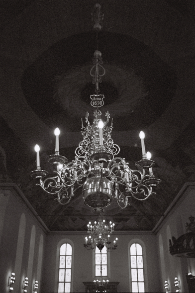 A grainy, black-and-white photo of a chandelier in Oslo Cathedral. The chandelier is not enormous, but it is very elaborate, with curly, organic-looking arms and decorations shining in the light from its now electric candles. At the top hangs a small shield saying ‘Anno 1698’. In the background is the cathedral's painted ceiling, some stained-glass windows and another chandelier on the other side of the nave.