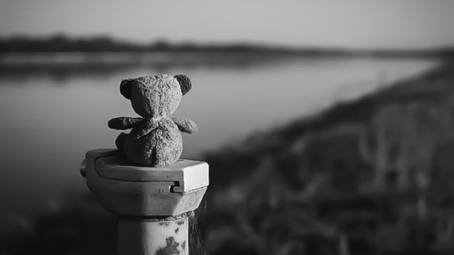 Ein kleiner Teddy sitzt auf einer Flußmarkierung und schaut auf den Rhein. Er ist dabei relativ gut zu sehen, während der Rhein unscharf im Hintergrund zu sehen ist. Das Foto ist in Schwarzweiß.