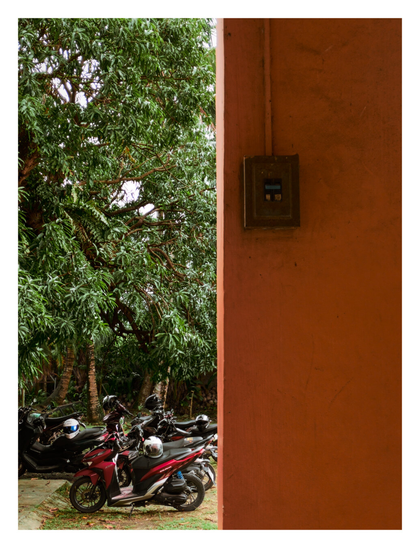 A large, textured orange wall or pillar on the right side, which features a mounted electrical box. To the left of the wall, a row of motorcycles and scooters is parked on a patch of grass and concrete. The vehicles, one of which is a distinct red and black, have helmets resting on them. The background is filled with dense, lush green trees. - Google Gemini 3 Preview
