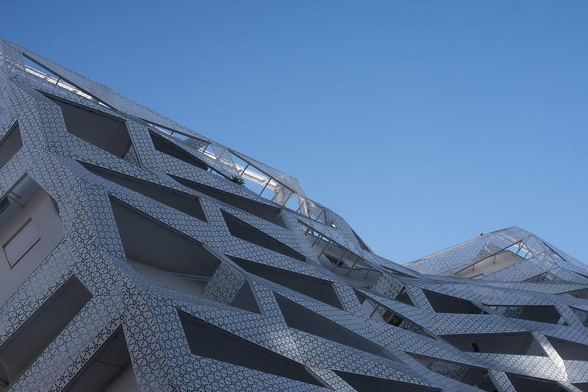 Photo en contre plongée d’un immeuble qui présente une architecture insolite, la façade n’étant pas plane, tout en angles, pleine de lignes brisée avec un matériau plein de motifs faisant comme de la dentelle d’acier