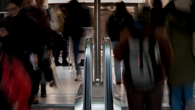 Foto vom Ende einer Rolltreppe, aufgenommen von der gegenüberliegenden Seite. In der Mitte sieht man ein senkrechtes Metallrohr, das den Zugang schützt, links und rechts daneben die Handläufe. Auf der linken und rechten Seite des Bildes sind verschwommen Menschen zu sehen, die die Rolltreppe nutzen.