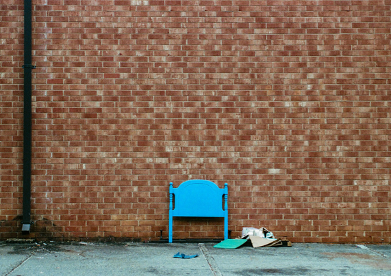 A photo of the back of a red brick building with a blue painted headboard/footboard from a bed leaning against it.