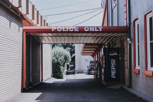 A covered driveway or alley between two buildings leads to a parking area, with a red and white "POLICE ONLY" sign mounted across the top of the passageway. The structure features a corrugated metal roof supported by red-trimmed posts. On the left side is a white-painted brick or siding wall, while the right side shows a building with gray siding and red trim. A Pepsi vending machine or sign is visible on the right wall. Through the passage, trees and parked vehicles can be seen in the bright sunlit area beyond. Overhead power lines cross the sky above the buildings.