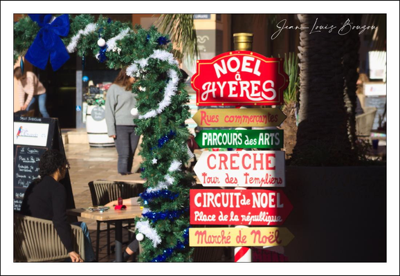 
A bright, hand-painted wooden signpost anchors the scene — a cheerful stack of red, green and white arrows nailed to a candy-cane-striped pole. The top plaque reads “NOËL A HYÈRES” in bold white letters; below are smaller directional boards with French phrases like “Rues commerçantes,” “Parcours des Arts,” “Crèche,” “Circuit de Noël,” and “Marché de Noël.” A thick evergreen garland, dotted with white lights and tied with big cobalt-blue bows, loops around the pole and frames the sign like a festive wreath.
Behind and around it the setting feels like a sunny, convivial street corner: a palm tree trunk rises beside the display, café tables and stacked chairs hint at nearby terraces, and a chalkboard menu leans in the background. People move through the scene out of focus, giving the image a lived-in, market-day energy. The whole composition reads warm and welcoming — a small-town holiday noticeboard catching the light and inviting passersby to stroll, shop and join the festivities.