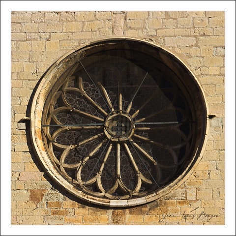 
A round, weathered rose window sits set into a warm, beige stone wall — the kind of circular tracery you expect on an old building. Radial stone spokes fan out from a small central roundel (which itself carries a simple cross-like motif), the spokes sweeping into elongated teardrop petals that create a flower-like pattern. A fine metal grille or mesh fills the openings behind the tracery, casting a delicate lattice of shadow across the interior and emphasizing the depth of the frame. The stone frame and mullions show age: chips, soft erosion and a mellow patina that catches the light and gives the whole piece a quiet, solemn dignity. The composition is perfectly centered and symmetrical, the geometry calming and slightly austere, while the interplay of light and shadow adds a touch of melancholy warmth. Overall it feels like a patient, enduring detail of an old building — both ornamental and functional, holding darkness and light in balance.