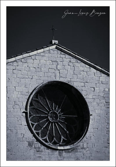 A stark, moody black-and-white photograph of the upper face of a small stone church. The triangular roofline slices into a deep gray sky, capped by a tiny cross that stands like a lone sentinel. Below, the wall is made of rough, square-cut stones whose uneven texture catches the light and casts small, crisper shadows.
Dominating the composition is a large circular rose window set into the masonry — a radial pattern of stone tracery that looks almost like a weathered wheel or the iris of an old eye. Deep shadows fill the window’s interior, creating a strong contrast with the pale facade and lending the whole scene a quiet, austere solemnity. A faint, hand-written signature sits in the upper corner, like a final, delicate flourish to the image. The overall effect is spare and contemplative: architecture rendered as silhouette and texture, simple lines and circles against a brooding sky.
