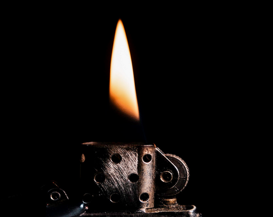 Man sieht den oberen Teil von einem Zippo-Feuerzeug. Dort sieht man die Flamme die durch dieses Feuerzeug entstanden ist. Dabei schaut es so aus, als wenn die Flamme über dem Feuerzeug schwebt.