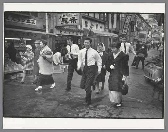 The image is a black and white photograph depicting several people walking down an urban street, indicative of daily life in Japan during the 1960s. Two men dressed in business attire are leading a group that includes women wearing traditional Japanese kimonos and modern coats or jackets, suggesting cultural diversity within this busy marketplace setting. The backdrop features storefront signs with Japanese characters, indicating various shops such as food vendors and possibly electronics stores.

One of the central figures is captured mid-stride, giving an impression of haste or urgency in their movement. There's a contrast between those who are walking together—perhaps colleagues—and individuals engaging separately, like the woman checking her items from a vendor stall. The street appears wet, hinting at recent rain and reflective surfaces that add depth to the scene.

The photograph is credited as "Mensen op straat in Osaka" by Ed van der Elsken, with an additional reference suggesting it was taken around 1960, aligning with historical records of urban life during Japan's post-war economic boom.