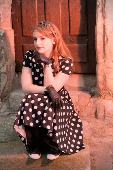 Une jeune femme rousse dans une robe noire à gros pois blanc, avec des gants noirs en cuir et un serre tête, assise sur des marches en pierre d’un petit escalier de 3 marches menant à une porte. Sa main gauche est juste en dessous de son menton, sa tête est tournée vers sa droite et ses yeux regardent la caméra