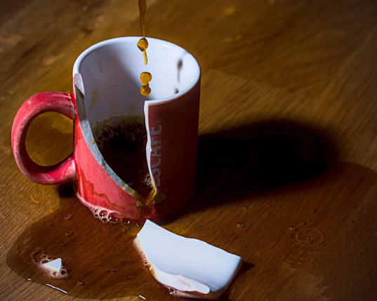 In eine Espressotasse fließt Kaffee. Die Tasse ist gesprungen. Ein großes Stück der Tasse liegt in der Pfütze des Kaffees, der aus der Tasse ausgebrochen ist. 