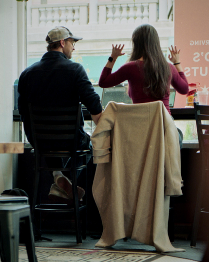 A man and a woman are seated at a cafe counter. The man, in a cap and dark jacket, faces forward, while the woman, with long hair and a maroon top, gestures with her hands raised. A beige coat hangs on her chair, and large windows open to a building view