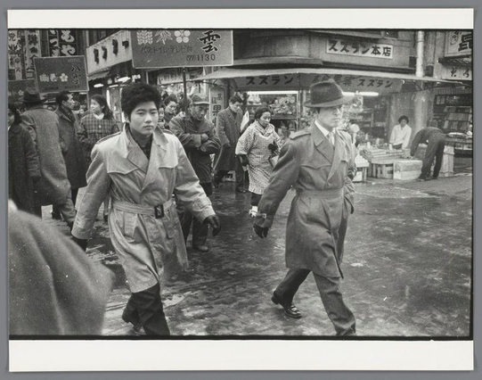 The image is a black and white photograph depicting two men in trench coats, walking briskly through what appears to be an urban street market. The man on the left has dark hair that falls just above his shoulders and wears glasses; he's wearing a light-colored trench coat with visible buttons and belt, as well as trousers tucked into boots. His companion is further ahead of him, sporting a hat and holding papers in one hand while carrying what seems to be a briefcase or bag on the other shoulder.

The street behind them is bustling with activity. Several individuals can be seen engaged in various activities: walking, shopping, some are interacting near market stalls that display goods for sale. The surrounding buildings have signs written in Japanese characters, suggesting this scene takes place in Japan, likely Osaka given the reference to Yakuza-mannen op straat in Osaka.

The pavement is wet and reflective, indicating recent rainfall or a cleaning activity. The overall atmosphere of the photo suggests it could be documenting an event from post-World War II era Japan with its depiction of everyday life amidst economic recovery efforts.

Additional information about this image can be found under Yakuza-mannen op straat in Osaka by Ed van der Elsken, dated 1960.