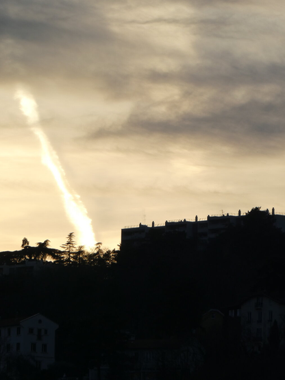 A contrail seeming to emanate from behind a hill, during sunset. The trail then goes behind a cloud.