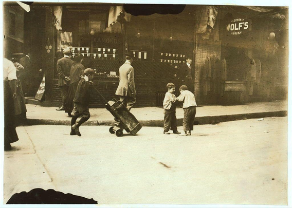 The image depicts a scene from the early 1900s on a bustling street in Boston, Massachusetts. The black and white photograph captures everyday life with three young boys as its main subjects. They are engaged in what appears to be an interaction or conversation while one of them is pushing a wooden cart along the sidewalk.

In the background, other individuals can be seen going about their day. There's a man walking away from the camera on his left side and another person partially visible at the far right edge of the frame. The street is lined with various establishments; notably "Wolf's House" clothing store in view to the top right corner.

The image carries historical significance, reflecting the social conditions and urban life during that era. It evokes a sense of nostalgia for early 20th-century America while shedding light on the innocence and resilience portrayed through these young boys' actions amidst their surroundings.