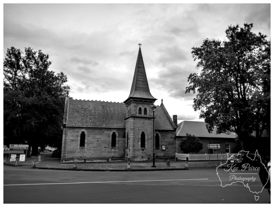 Black and white photograph of the historic Ross Uniting Church in Tasmania, featuring its central stone tower and spire. 

The sandstone building is set at a street corner, framed by large, dark trees on the left and right, with a white picket fence visible on the right. The sky is heavily clouded.