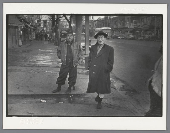 The image is a black and white photograph showing two men standing on the sidewalk in what appears to be an urban street setting. The man on the left is wearing casual attire, including a jacket with prominent arm stripes, possibly indicating some form of affiliation or membership, along with dark trousers and boots. He has his hands clasped behind his back and stands slightly leaning against a pole for support.

The man on the right exudes an aura of authority; he's dressed in more formal clothing—a long coat with buttons down the front, knee-length socks paired with shoes that seem to be polished leather—complementing this ensemble is a wide-brimmed hat. He has his hands casually tucked into his pockets and stands upright.

The street behind them features various storefronts, signage indicating businesses or shops in Japanese characters, suggesting an East Asian setting. There's also a hint of vehicular traffic with what appears to be the front end of a car visible at the far right edge of the photo.

There is no discernible movement from either individual; they both seem still and attentive as if posing for the photograph. The ground shows wet patches, indicating recent rain or damp conditions on the street surface. A subtle detail in this candid snapshot captures an everyday moment with potential stories behind it—perhaps these men are local characters within a neighborhood narrative.

The image is labeled "Elsken" a [...]