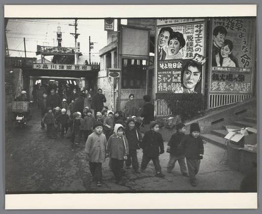 The image depicts a bustling street scene in what appears to be an urban area, possibly Japan during the early 1960s. A group of children is walking along the sidewalk towards and away from the camera's perspective, with some adults accompanying them. The expressions on their faces suggest curiosity or engagement as they navigate through this busy environment.

The street is lined with various signs in Japanese script, advertising food items like tofu and noodles, which indicates a focus on local cuisine culture at that time. There are also large banners featuring images of people's faces, likely advertisements for movies or public events, adding to the sense of an active community life. In one instance, there seems to be a vendor with goods displayed near the street.

The architecture is relatively simple and functional, typical of mid-20th-century urban development in Japan. There are visible elements like power lines strung overhead and various forms of commercial signage that contribute to the busy atmosphere captured by Ed van der Elsken's camera lens.

This photograph likely serves as a historical record or artistic portrayal of daily life for children amidst cultural influences, reflecting societal changes during an era marked by growth in urbanization.