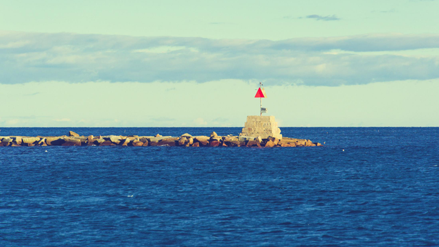 A short obelisk with a triangular sign and probably a light is on the edge of a thine jetty of light colored boulders just below the horizon of a slightly choppy sea with languid clouds stretched horizontally above
