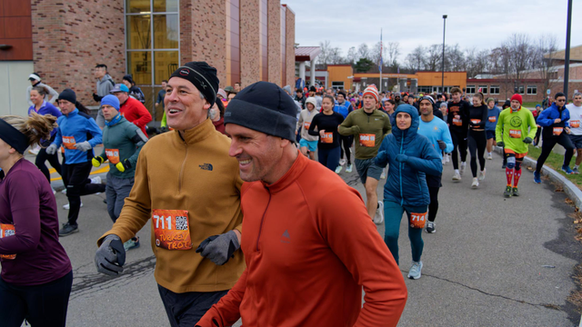 Two men in two different shades of orange jacket and green caps seem to be running together as friends in a large crowd of other runners coming from a high school entrance in the distance.