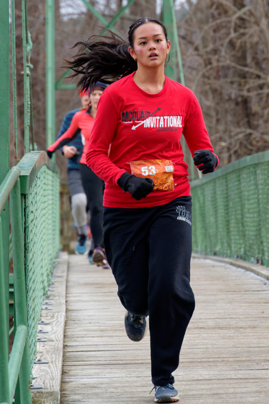Woman wearing a red shirt and black pants is running with her black hair in a ponytail splayed to the the left with bib number 53 and black gloves.  She's running on a bridge with a floor of fine wooden horizontal slats with green fence sections and suspension cables above